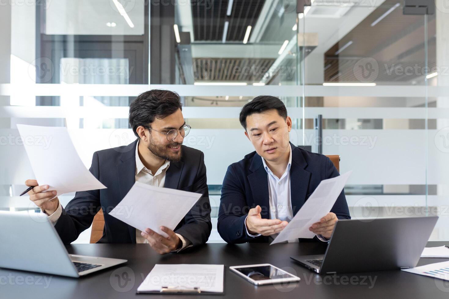 Two business professionals engaging in discussion while reviewing important documents. Workspace includes laptops, tablet, and papers. Office setting promotes teamwork, collaboration photo