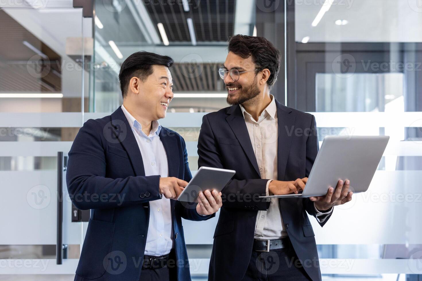 Confident business professionals engaged in friendly conversation using tablet and laptop. Image captures modern workplace, teamwork, and professional attire, highlighting collaboration photo