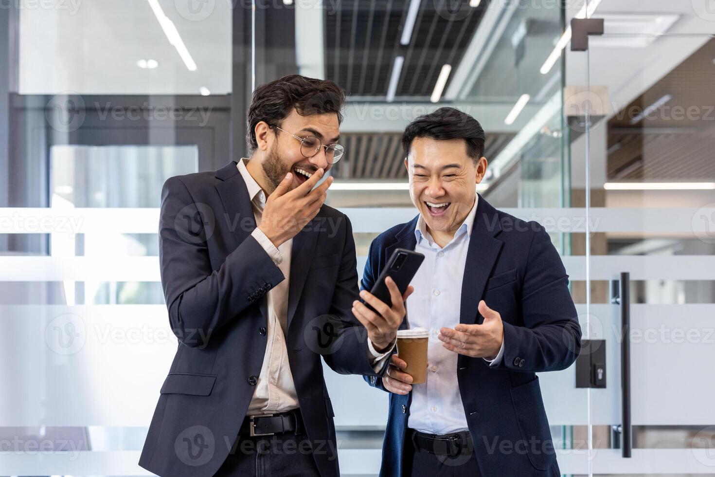 Two colleagues sharing a light moment in office while watching a funny on smartphone. Both men dressed in business suits, one holding a coffee cup, expressing laughter and surprise. photo
