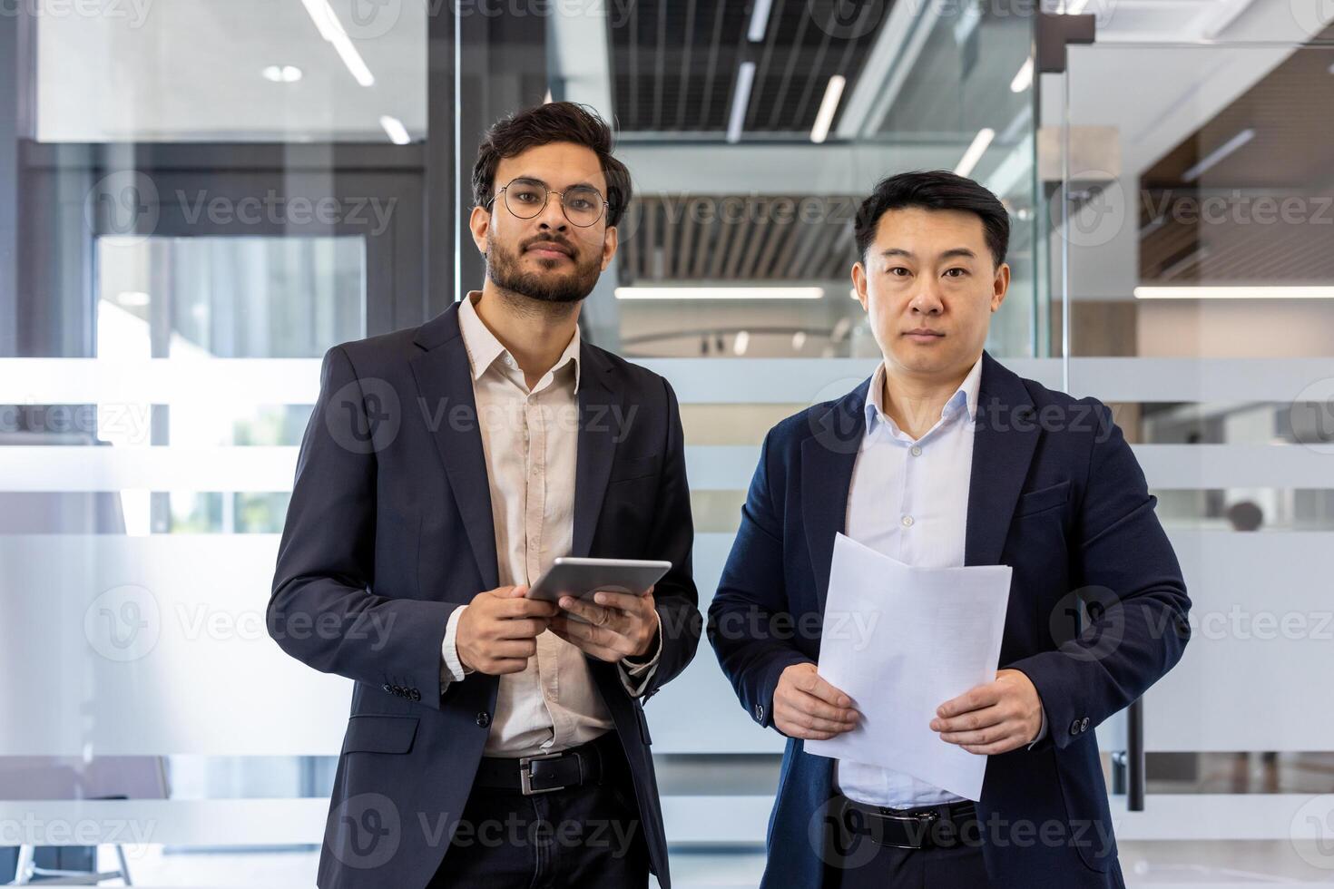 Two business professionals standing in modern office holding documents and tablet. Serious expressions indicate focus on business project. Office setting suggests teamwork and professionalism. photo