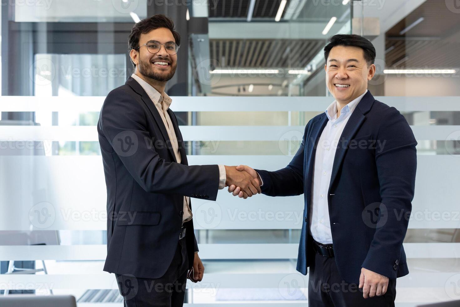 Two businessmen in suits exchanging a confident handshake in modern office. They are smiling, indicating successful collaboration or partnership. Image captures concepts of business success photo