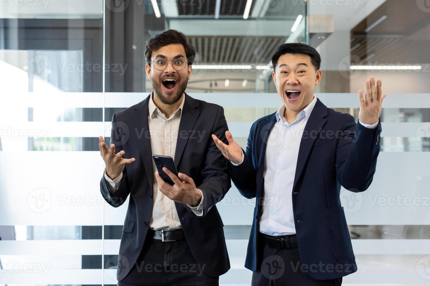 Two businessmen in formal attire expressing excitement and surprise in modern office environment. One holds smartphone, emphasizing technology and achievement. Perfect representation of teamwork photo