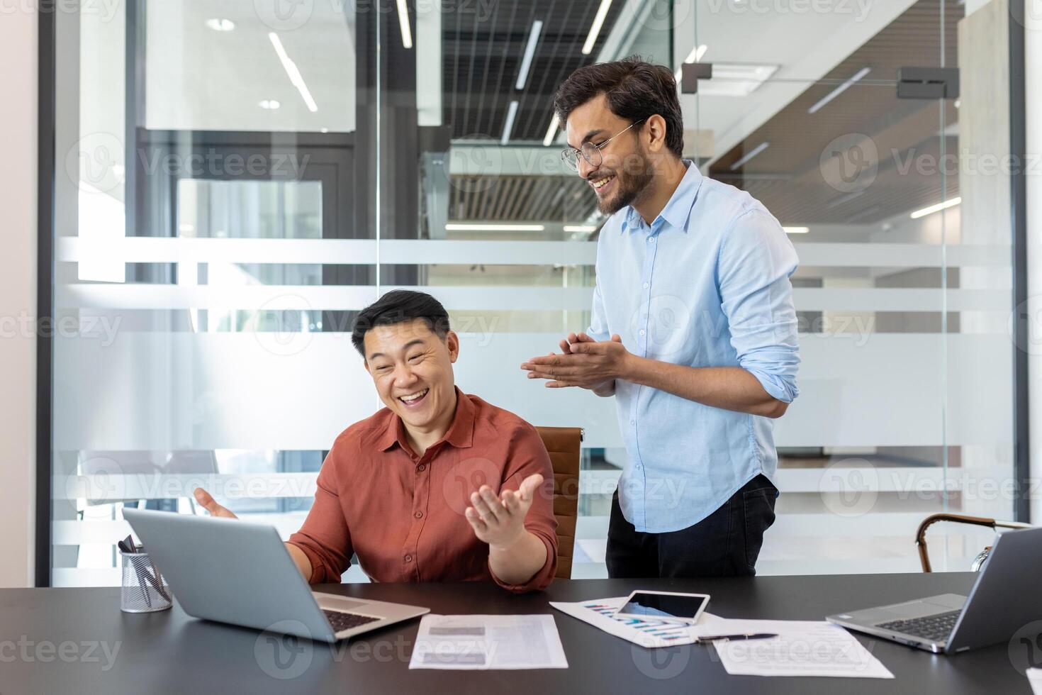 Two coworkers are seen in bright, modern office setting, energetically discussing and celebrating work success. The table displays graphs and a smartphone, emphasizing a productive workplace ambiance photo