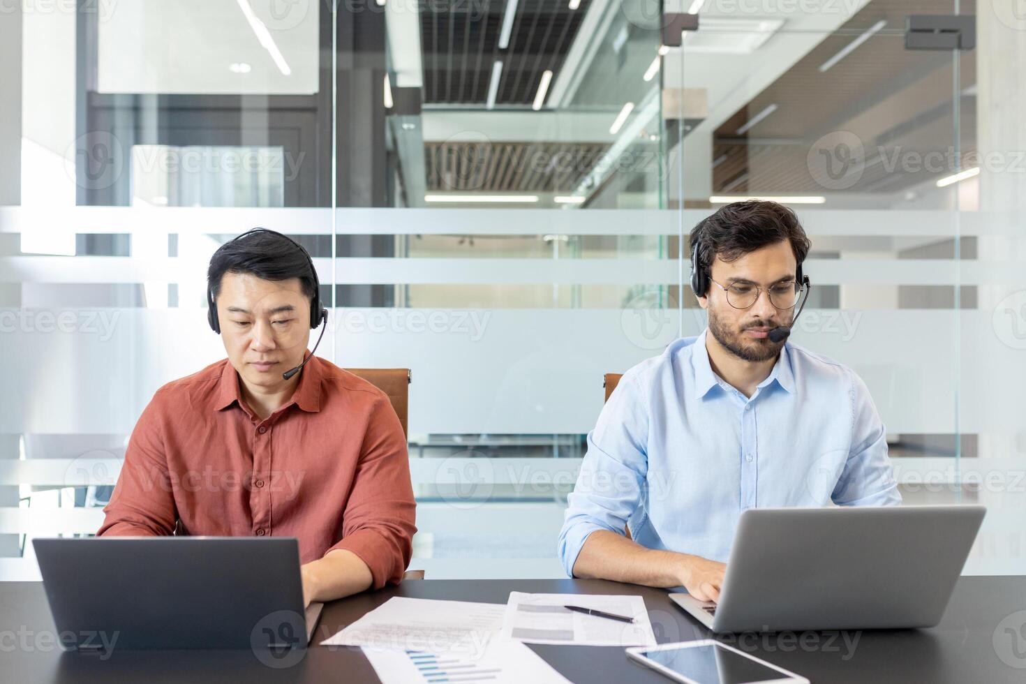 Two colleagues in casual clothes working inside office with laptops and headset. Office workers consulting clients remotely while sitting at desk. photo