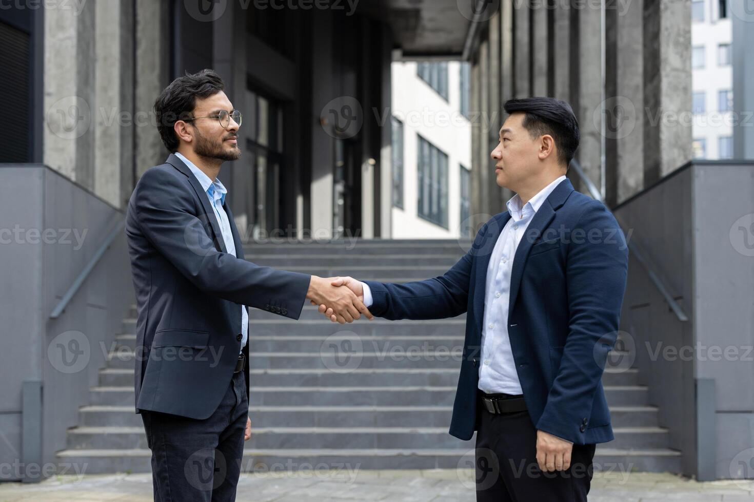 Two serious men in business suits greet each other shaking hands outside an office building. Diverse business people walking through the city. photo