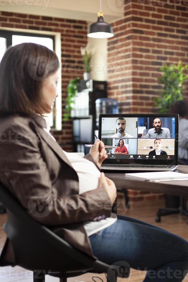 Selective focus of laptop displaying group conference call as pregnant businesswoman listens to her coworkers. Expectant lady with hand on pregnancy belly having videocall on digital device at office. photo