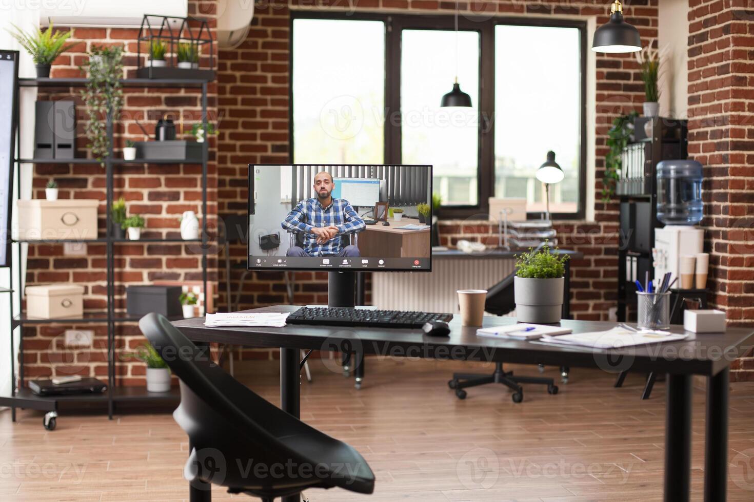 Vacant brick wall workplace setup with computer screen showing videocall of businessman in wheelchair. Desktop pc displaying ongoing virtual meeting with caucasian man in empty startup office. photo