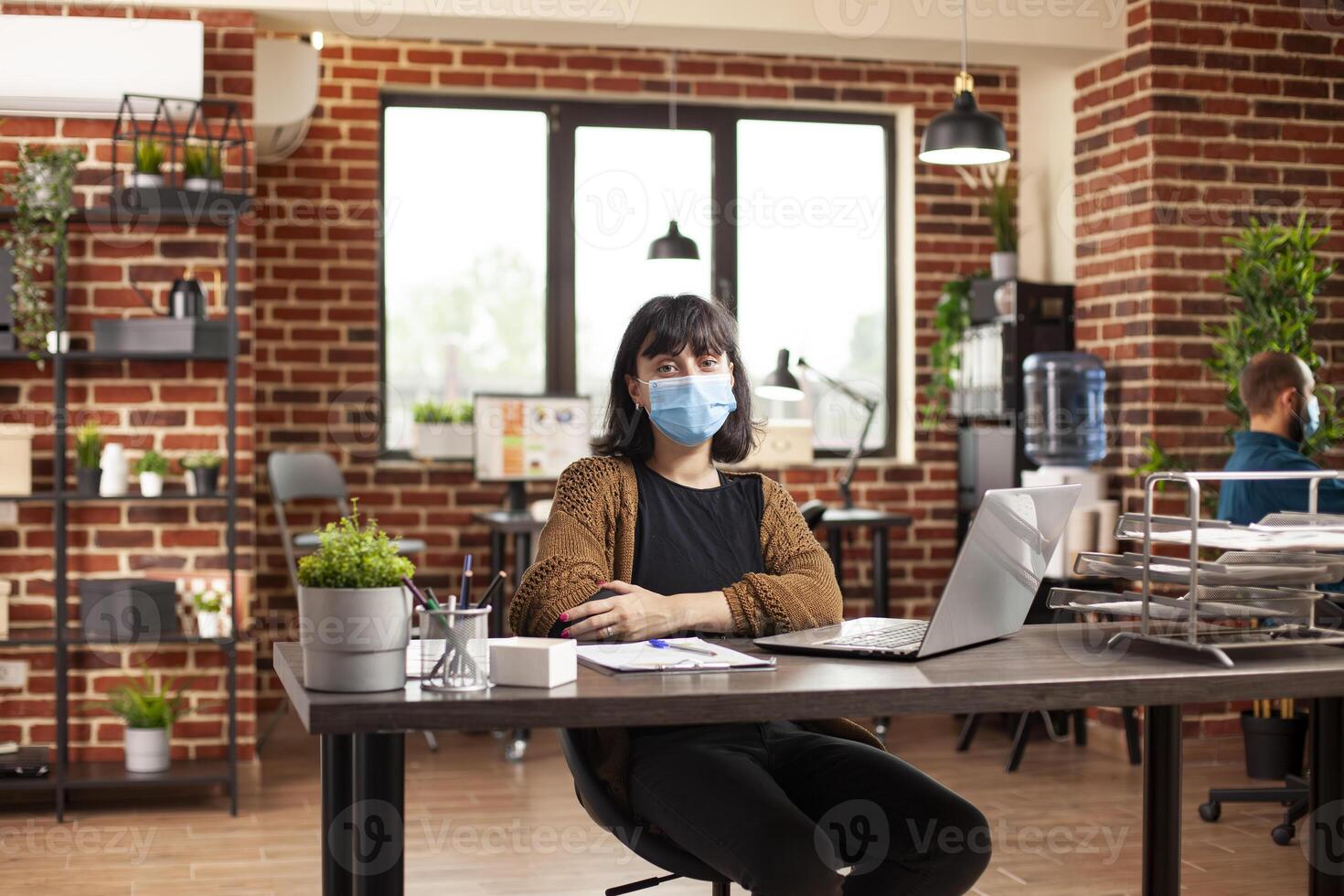 Young caucasian woman poses confidently at desk arranged with laptop and financial documents in startup office. Female employee wearing face mask and looking at camera in brick wall workspace. photo