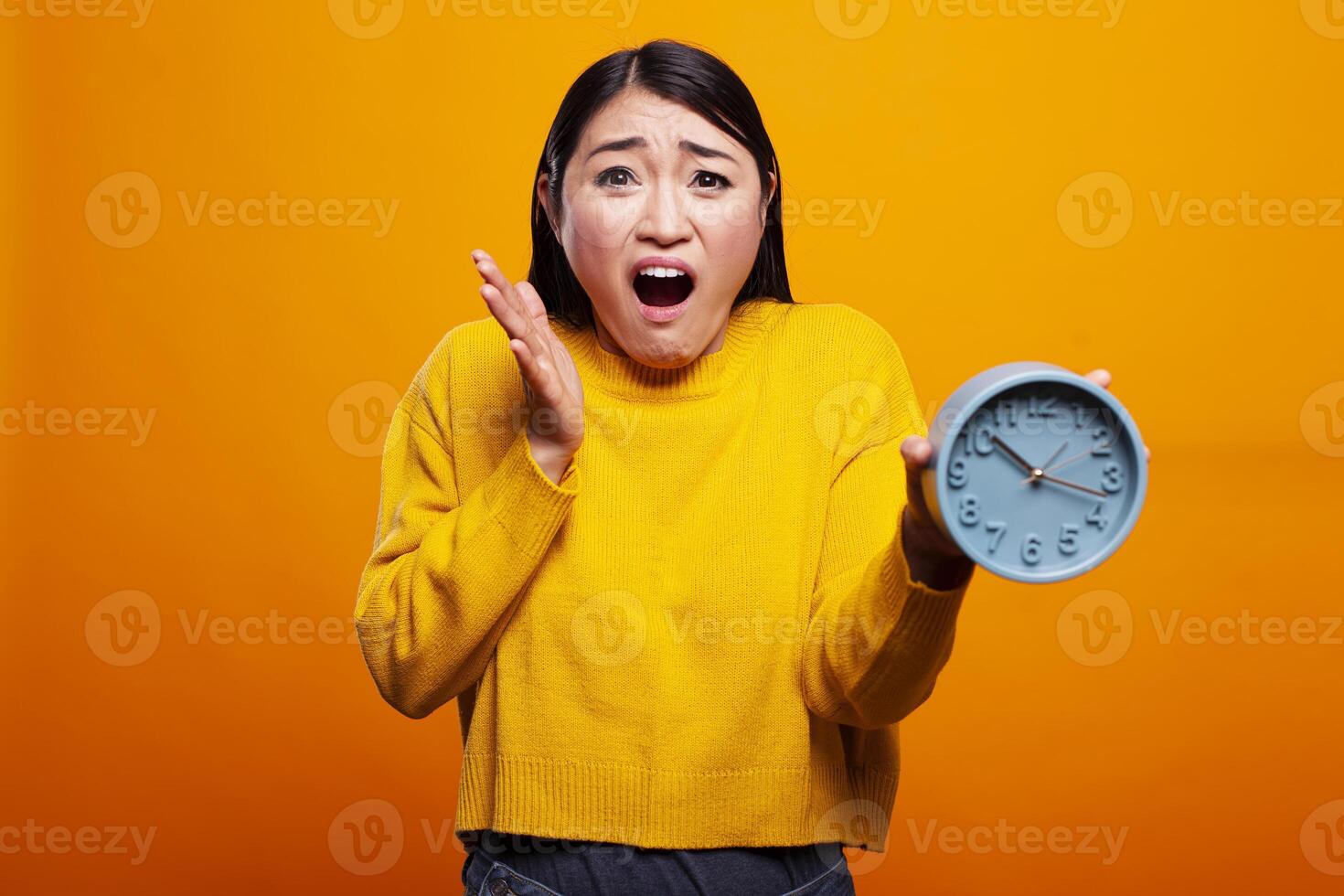 Asian woman in yellow sweater holds a blue clock with shocked expression, showcasing urgency and stress of managing time. Female individual looking surprised and worried, running late for appointment. photo