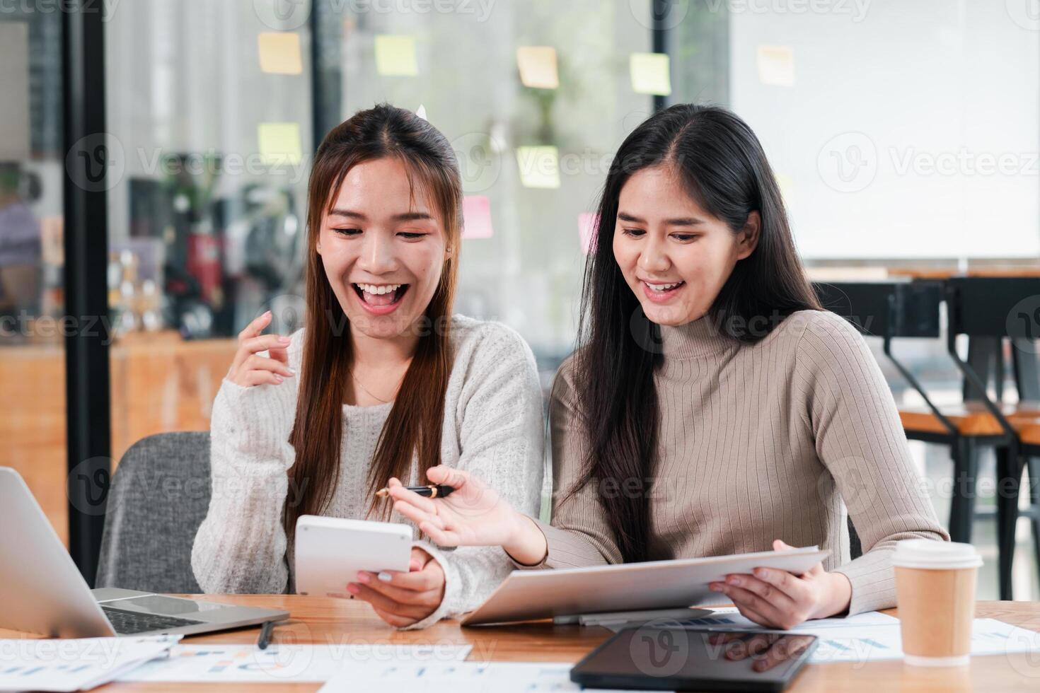 Two young women in a modern office, collaborating with laptops and documents, smiling and engaged in creative work. photo