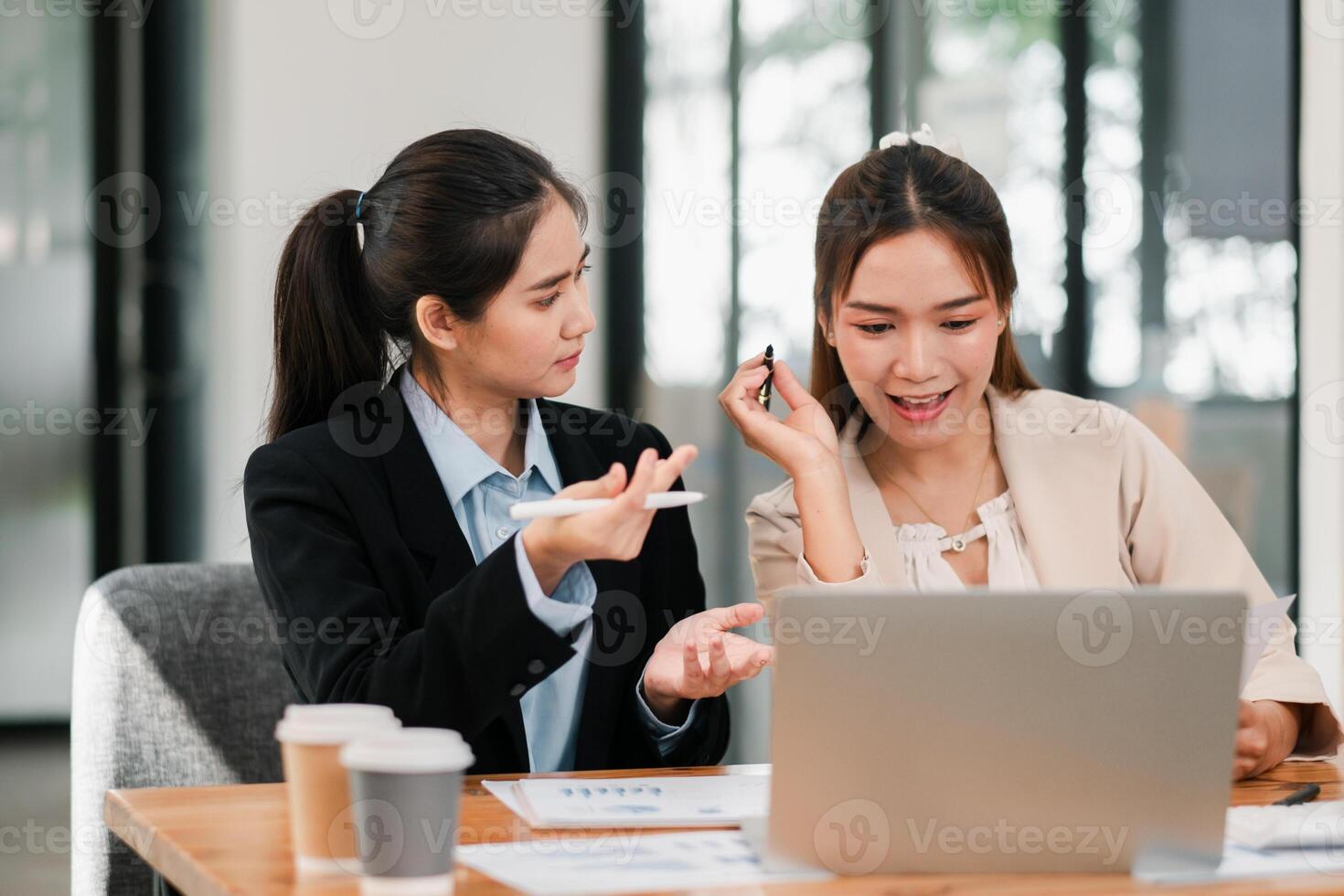 Two businesswomen discussing work at a desk with a laptop and coffee cups in a bright, modern office environment. photo
