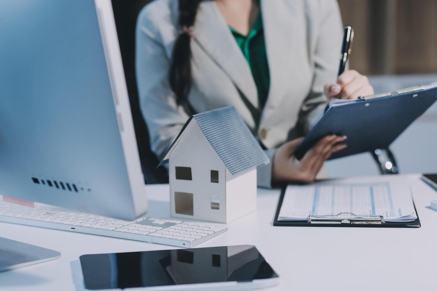 A woman is writing on a clipboard while sitting at a desk with a house model photo