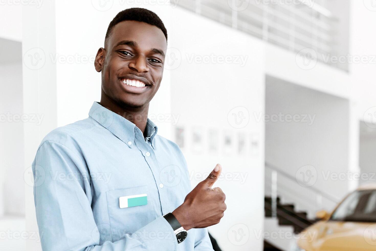 Cheerful black car salesman showing thumb up gesture, recommending best automobile dealership, copy space. Positive Afro seller working with laptop pc, looking at camera in auto showroom photo