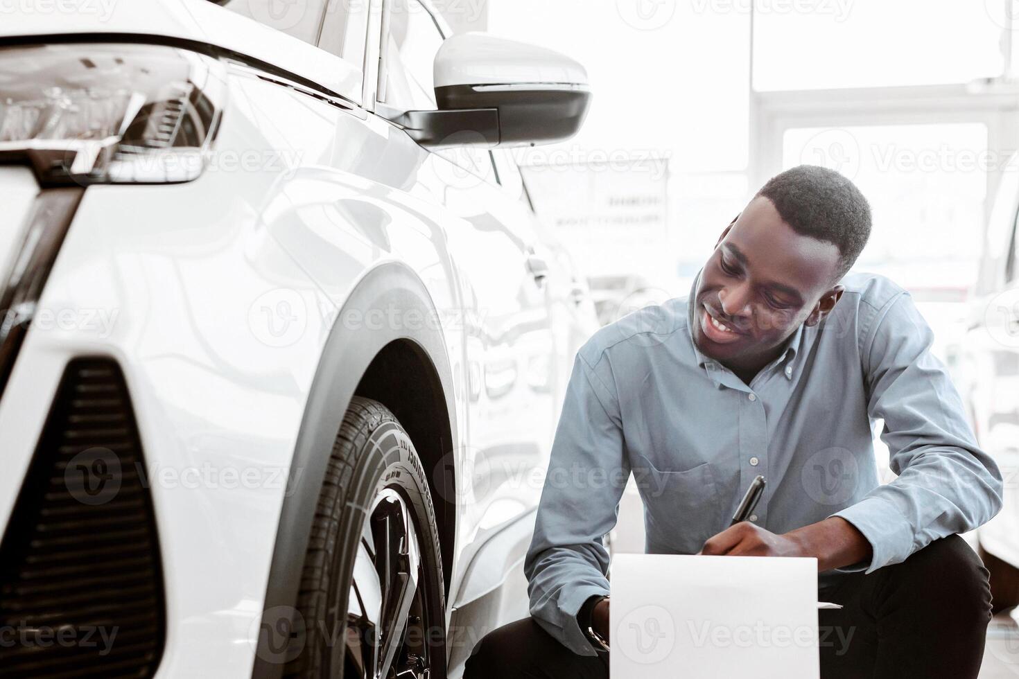Smiling black car salesman sitting near new auto, writing in clipboard at contemporary automobile dealership. Professional manager working at vehicle showroom, copy space photo