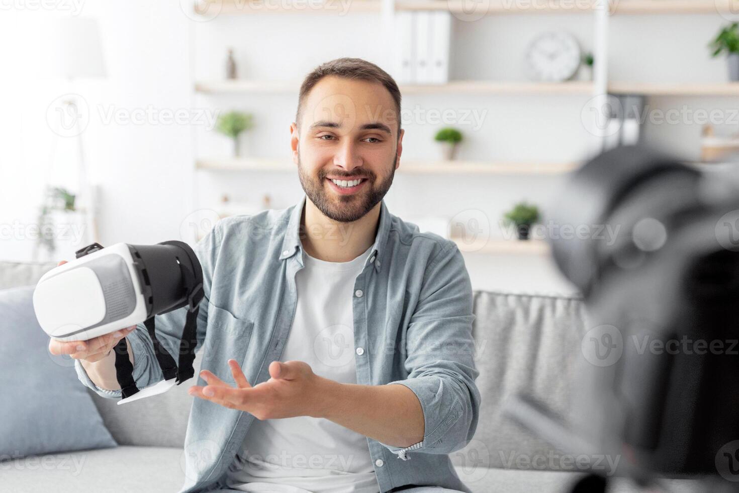 Young male blogger shooting review of virtual reality headset, using professional camera at home. Millennial content maker promoting modern gadget, filming for social media photo