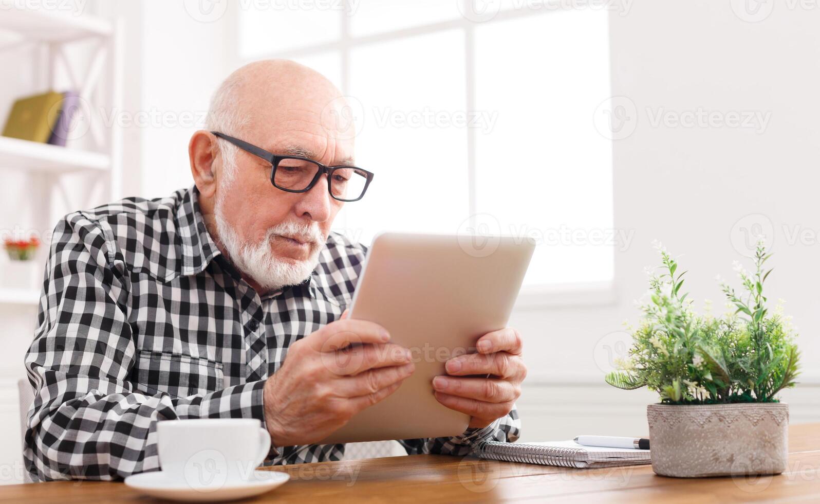 Senior man reading news on digital tablet. Serious mature male using portable computer at home, sitting at table, copy space photo
