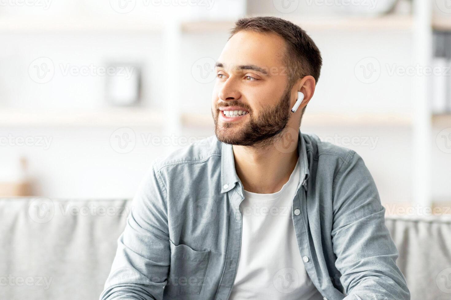Smiling young man in earphones using laptop for online communication at home office. Millennial Caucasian freelancer having call, chatting remotely via computer, indoors photo