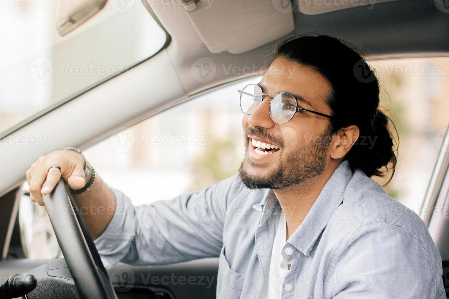 Joyful long-haired indian guy with glasses taxi diver driving brand new car, looking at rear view mirror and smiling, side view, copy space. Cheerful middle-eastern young man enjoying his new auto photo