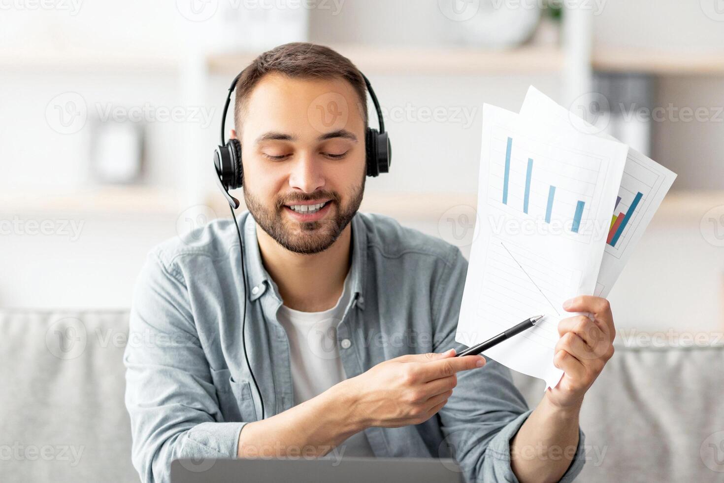 Cheery young guy in headset making call, showing documents at webcam, using laptop, working from home. Millennial man communicating online, participating in web conference or business meeting photo