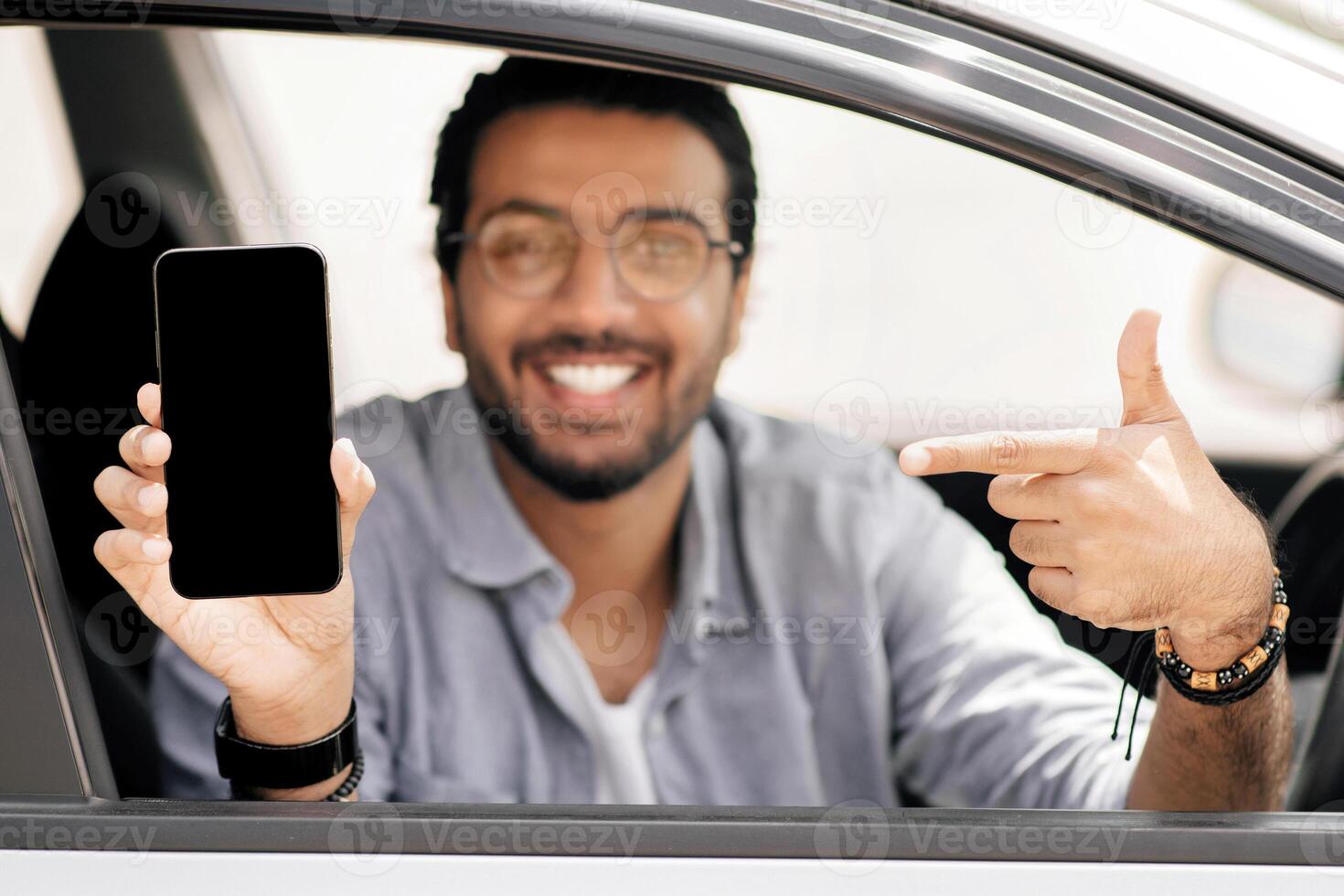 Selective focus on mobile phone with empty display in smiling middle-eastern guy hand, arab man driver using mobile application for navigation or car renting service, copy space, mockup photo