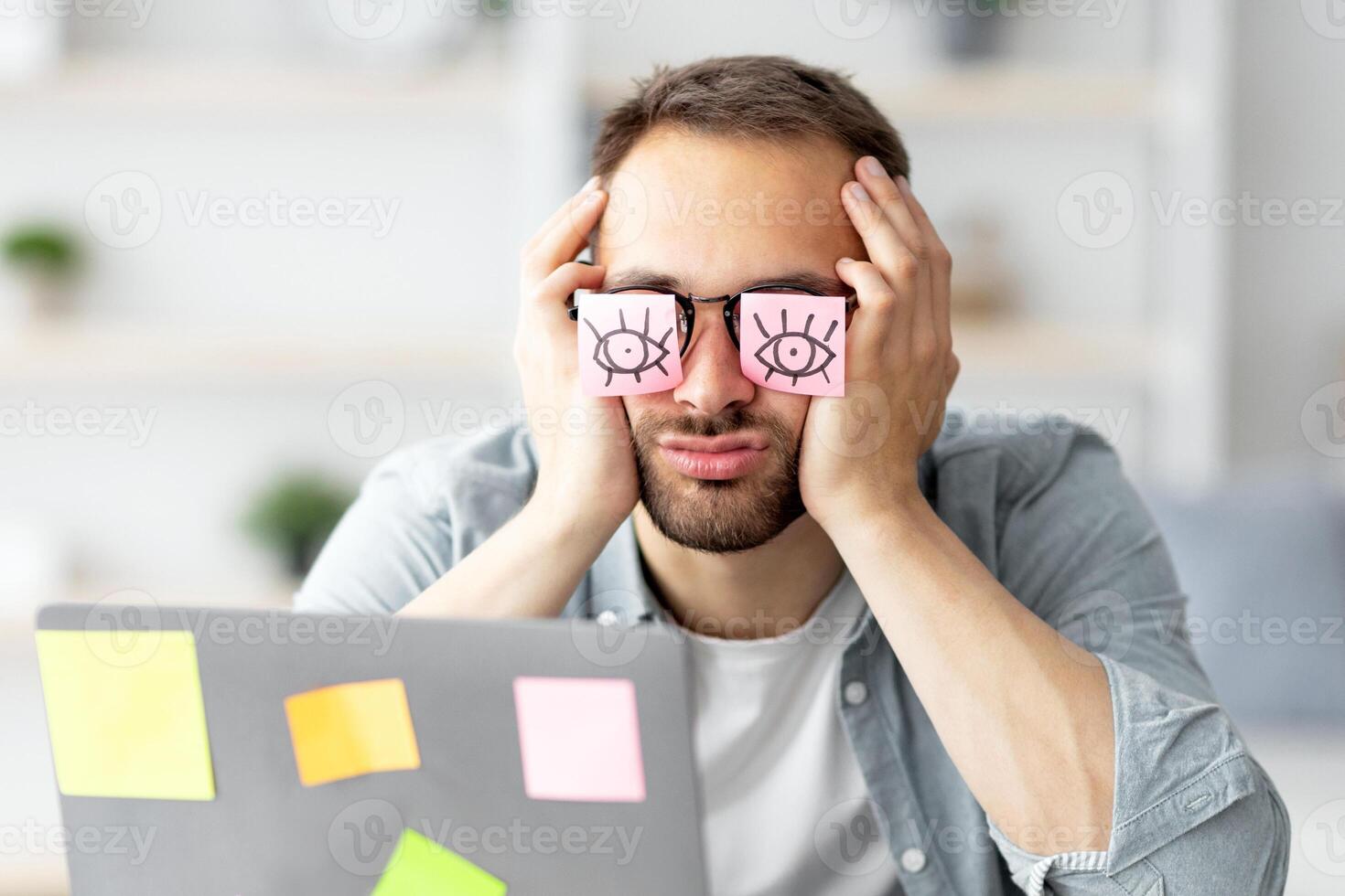 Tired young man having stickers with open eyes drawing on his glasses, sleeping in front of laptop at home office. Exhausted millennial man overworking, napping, failing to meet deadline photo