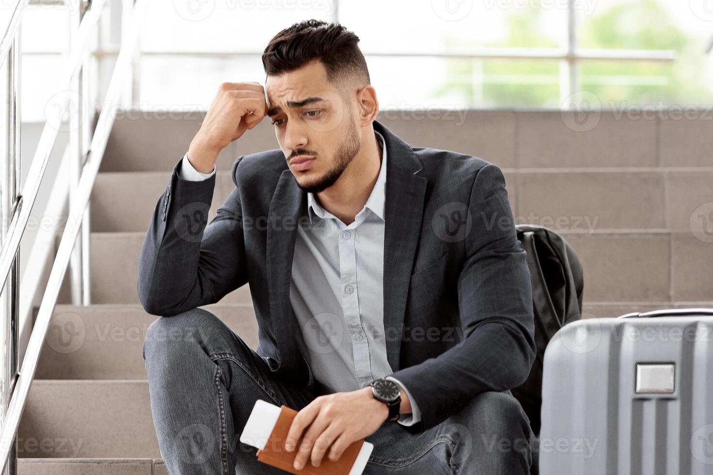 Upset Middle Eastern Guy Sitting On Airport Terminal Stairs With Luggage And Passport In Hands, Depressed Young Arab Man Missed His Flight, Touching Forehead And Thinking About Problem Solution photo