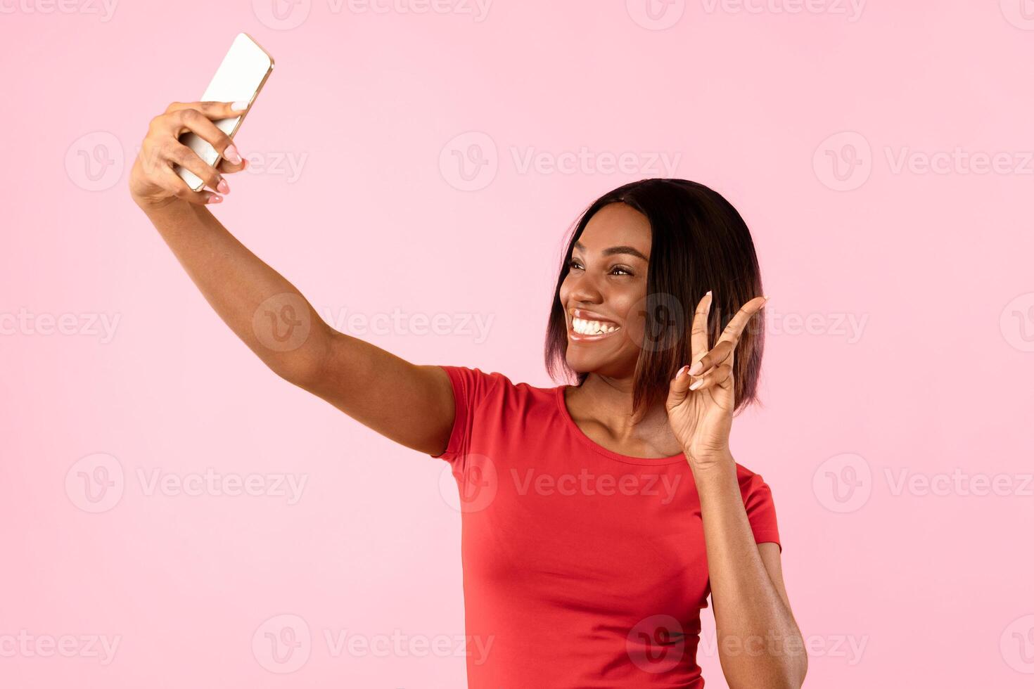 Cheerful African American Lady Making Selfie On Mobile Phone Posing Gesturing Victory Sign Over Yellow Background In Studio. People Having Fun Using Gadgets. Social Media, Blogging Concept photo