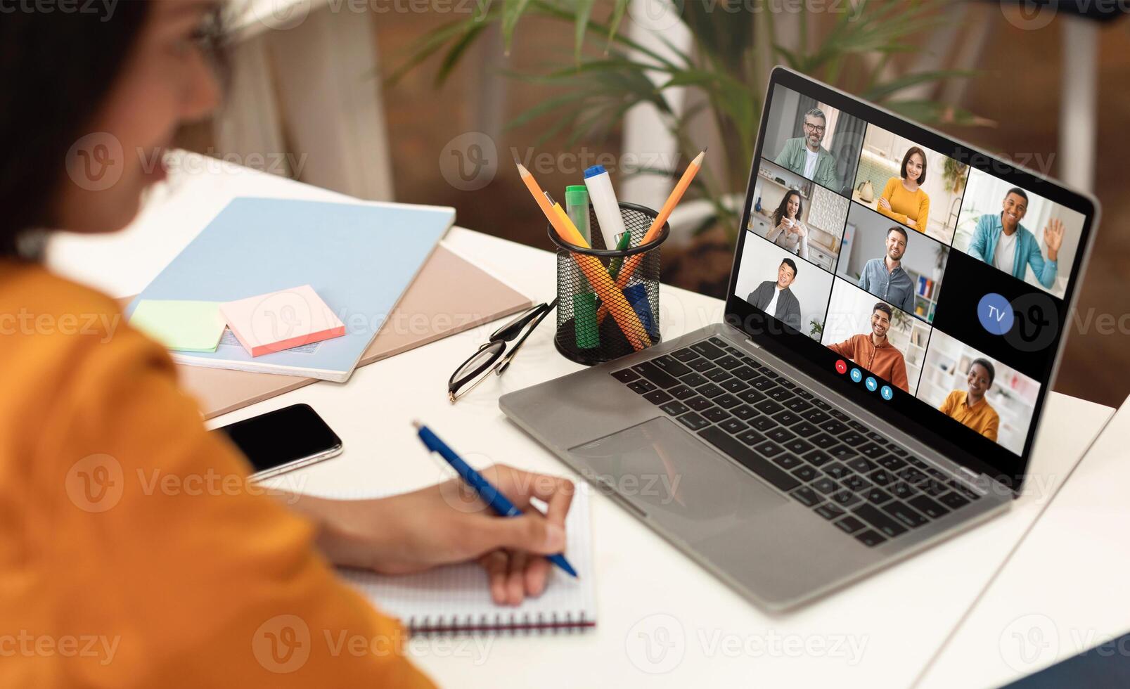 A woman sits at a desk with a laptop open, displaying a conference meeting with several participants. She is taking notes in a notebook with a blue pen, her focus on the screen. photo