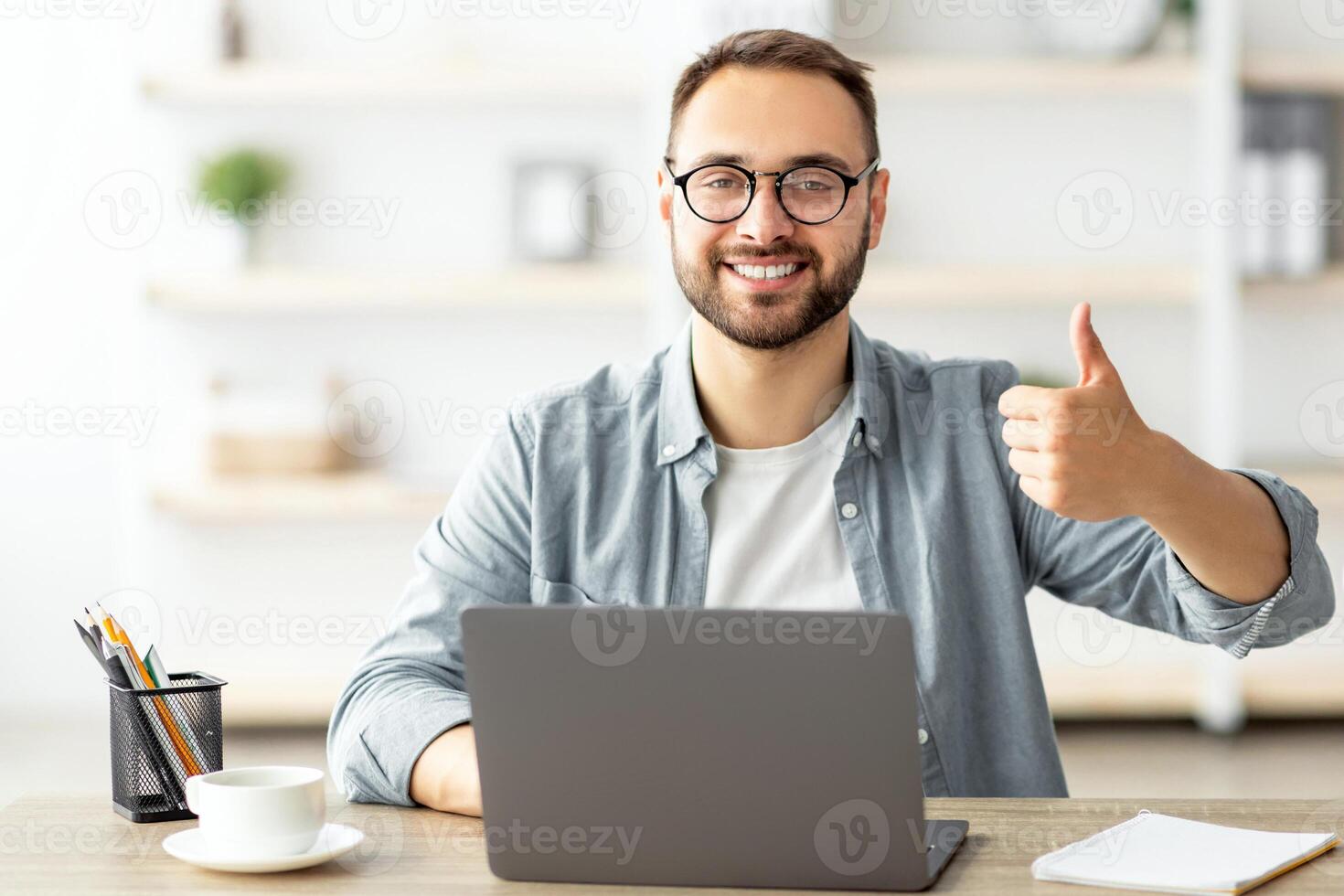 Happy young guy sitting at table with flag of Canada, using laptop computer, showing thumb up gesture at home. Modern online foreign education, emigration and citizenship, working abroad concept photo