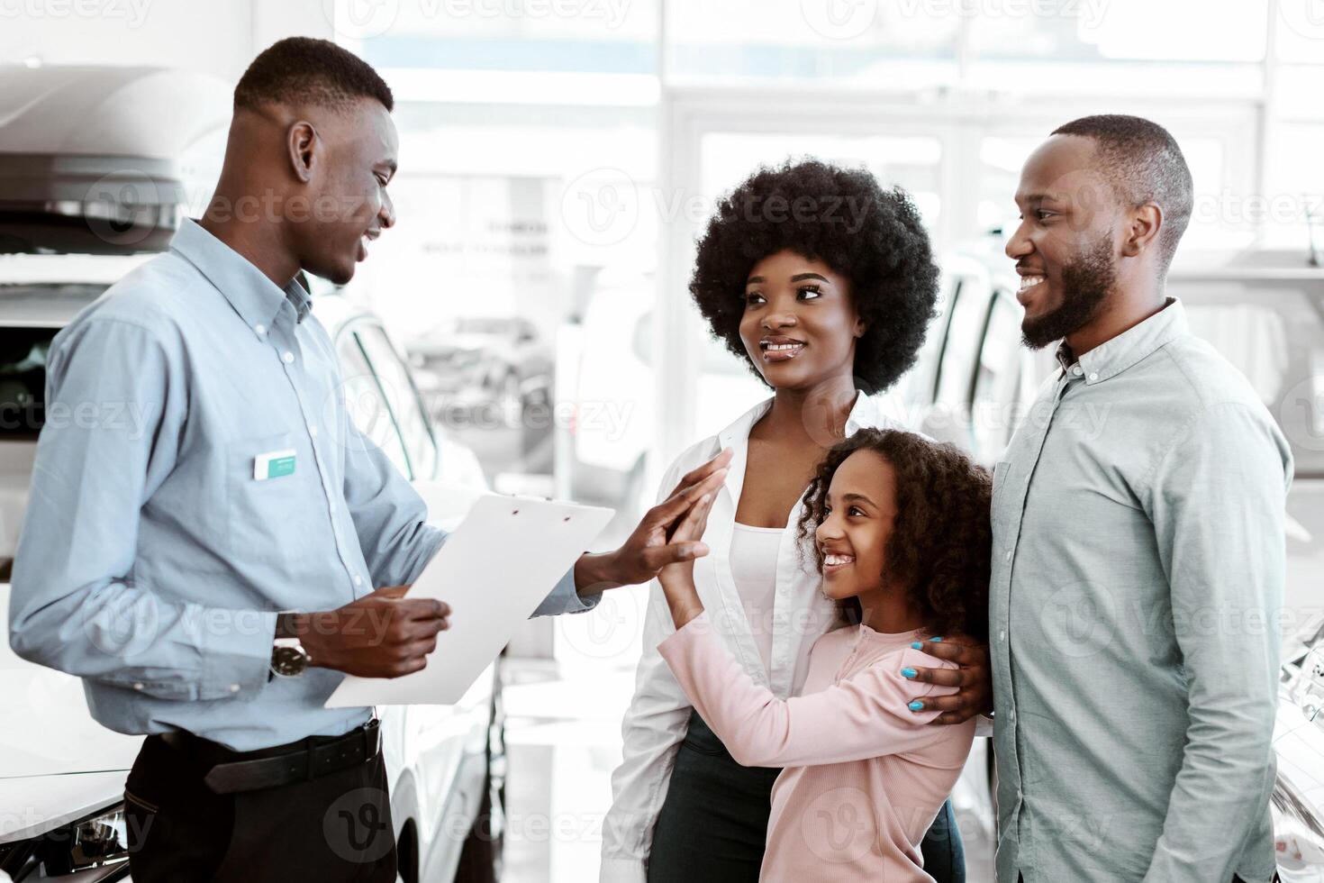 Cute Afro girl high fiving positive salesman at car dealership. Friendly black family purchasing or renting new car, making agreement with seller at automobile showroom photo