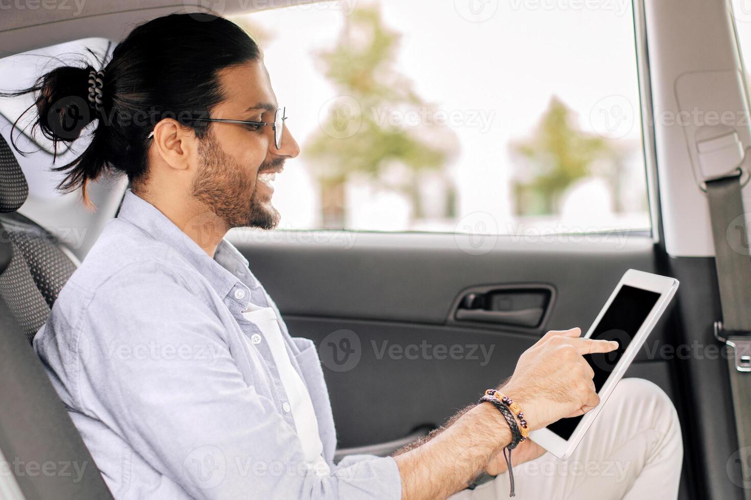 Side view of arab guy in casual outfit sitting at taxi, using pad, copy space. Cheerful middle-eastern young man using transportation service, sitting at automobile back seat, surfing on Internet photo