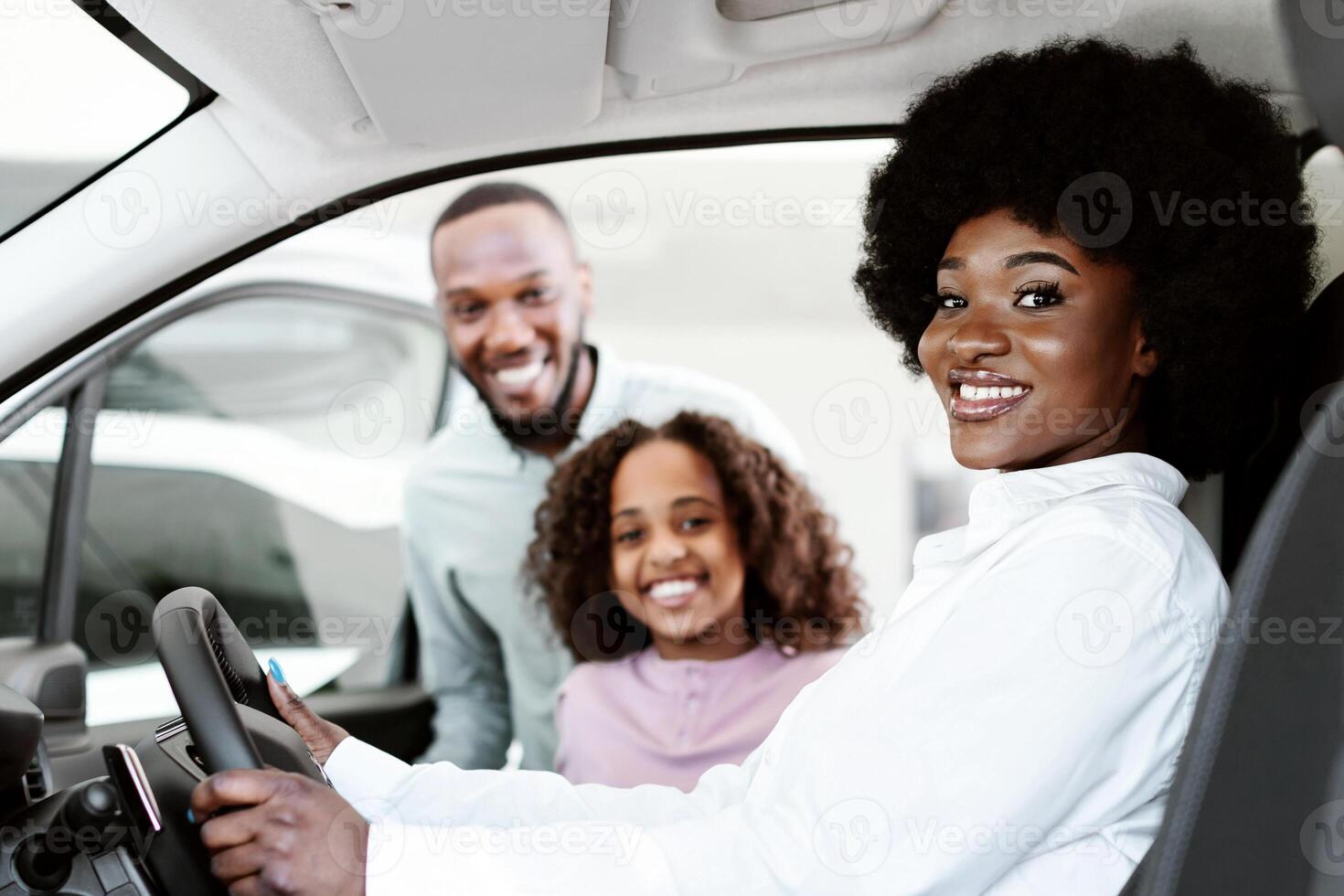 Test drive. Happy black woman sitting in driver seat of new car, choosing vehicle with husband and daughter at auto dealership. Afro family checking automobile before purchase or rental photo