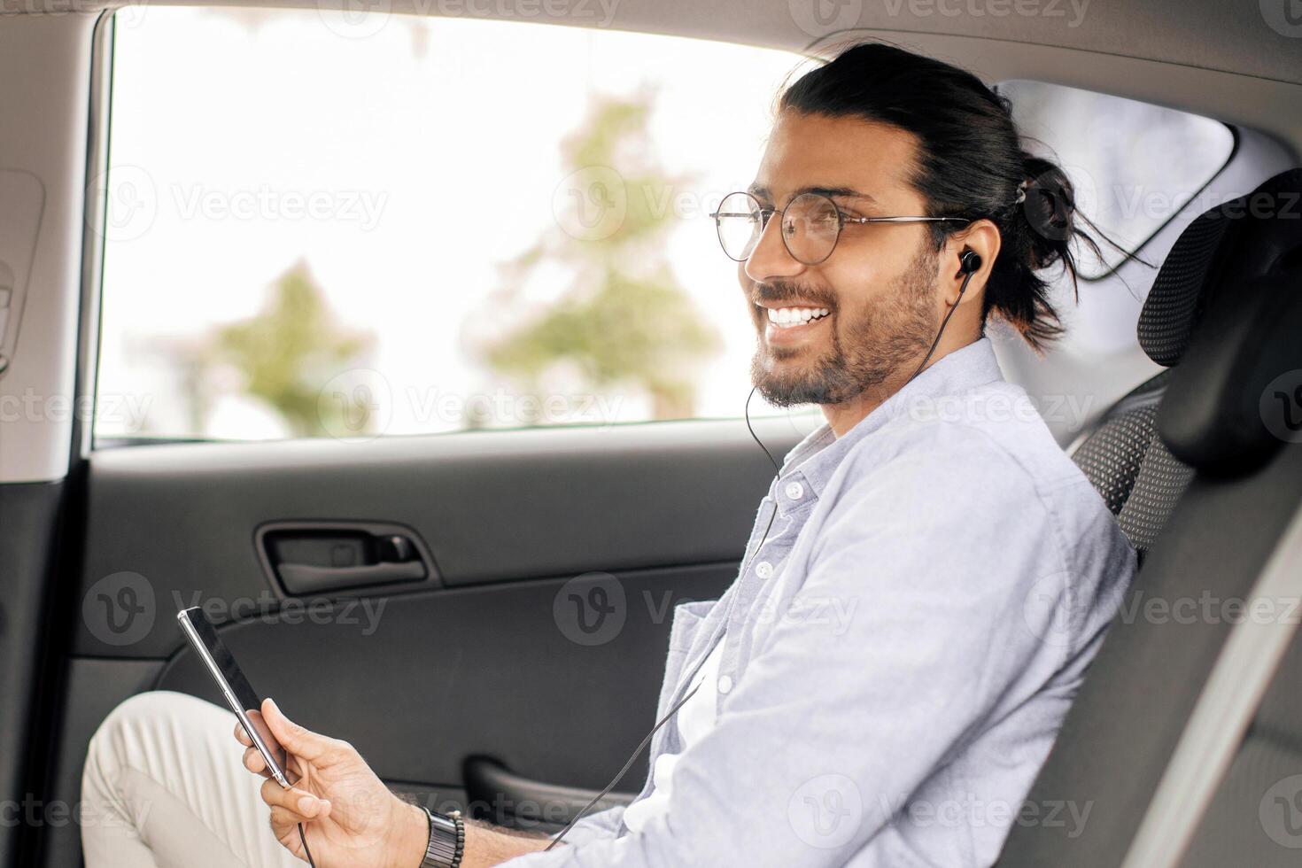 Cheerful middle-eastern man sitting at auto back seat, listening to music while going somewhere buy transportation service, using earphones and mobile application on smartphone, copy space photo