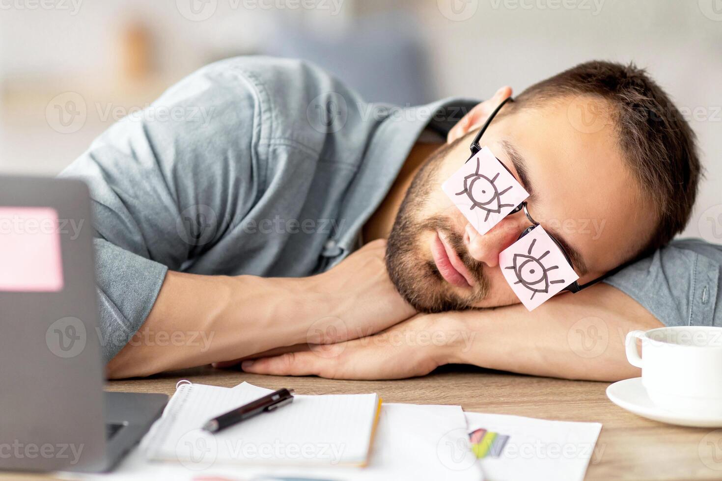 Lazy unproductive young guy wearing funny sticky notes with open eyes on his glasses, sleeping at workplace. Tired overworking millennial man falling asleep at home office photo