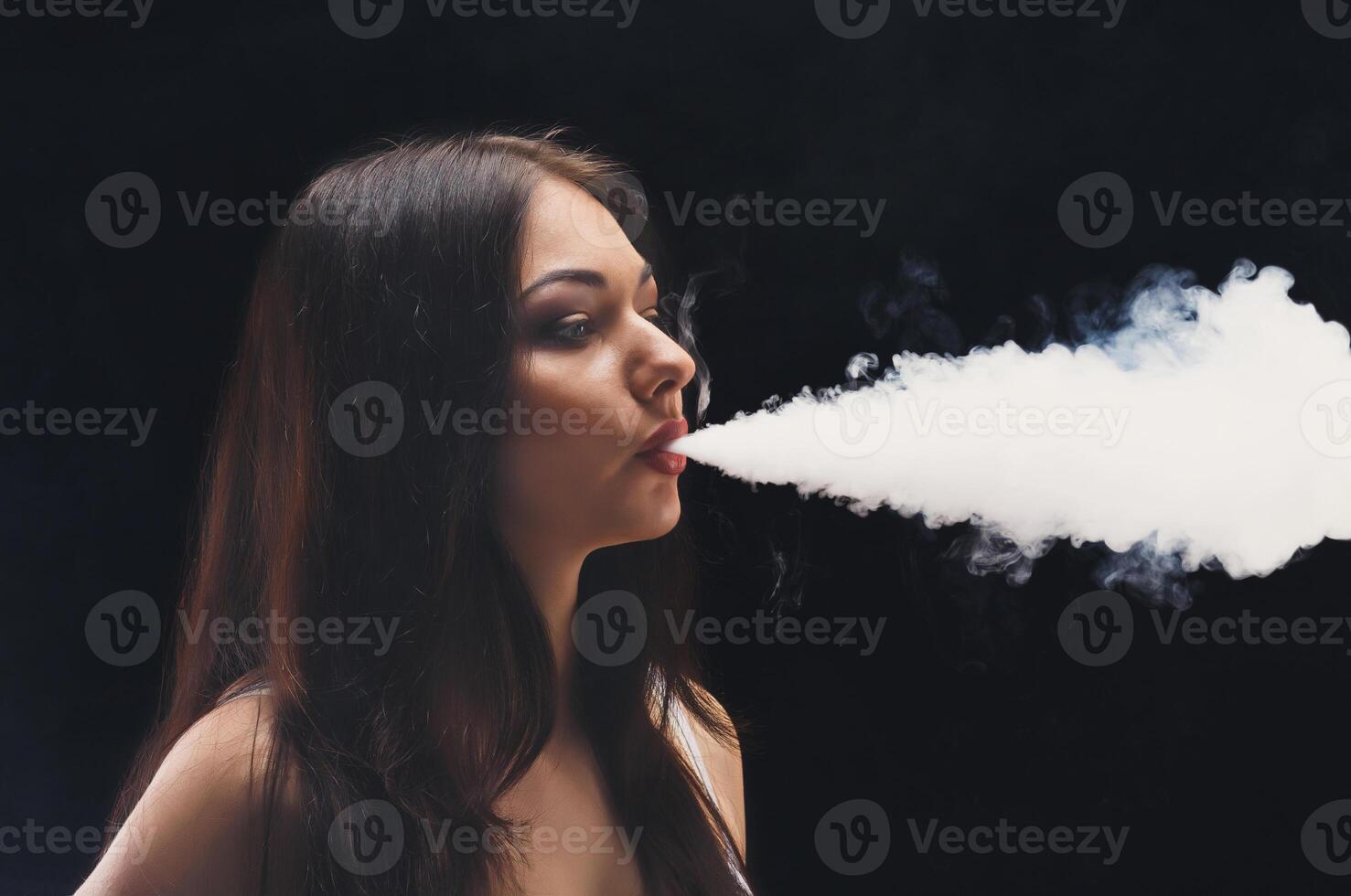 Brunette woman vaping electronic cigarette with smoke on black background closeup. Young woman smoking e-cigarette to quit tobacco. Vapor and alternative nicotine free smoking concept, copy space photo