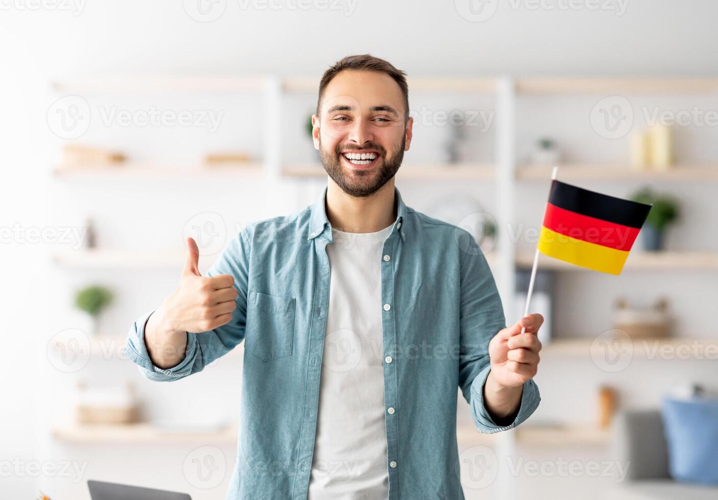 Happy Caucasian man showing thumb up and flag of Germany, posing and smiling at camera indoors. Cheerful millennial student recommending foreign education, learning German language photo