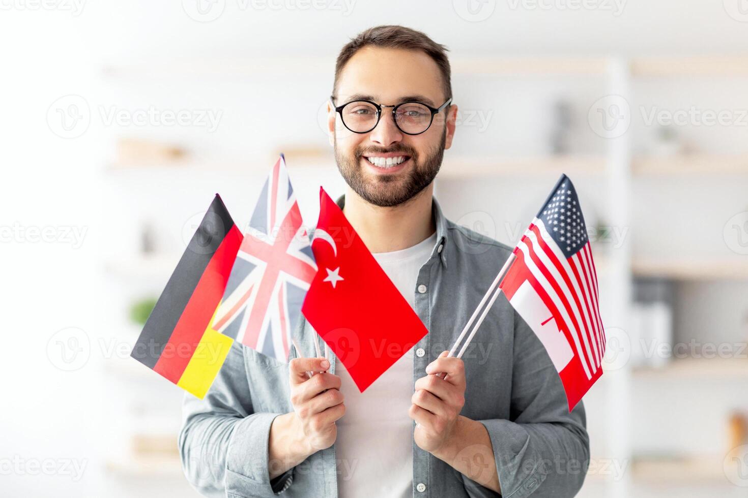 Handsome young guy holding bunch of diverse flags and smiling at camera indoors. Happy millennial man recommending foreign languages school, emigrating abroad. Modern education and student exchange photo