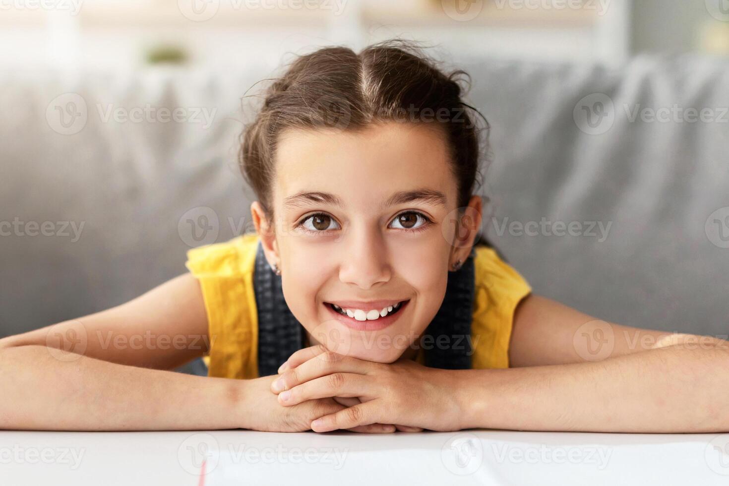 Happy Kid. Front Headshot Of Cheerful Adolescent Schoolgirl Posing And Looking At Camera, Little Female Sitting On Sofa Leaning On Table And Hands. Positive Facial Expression photo