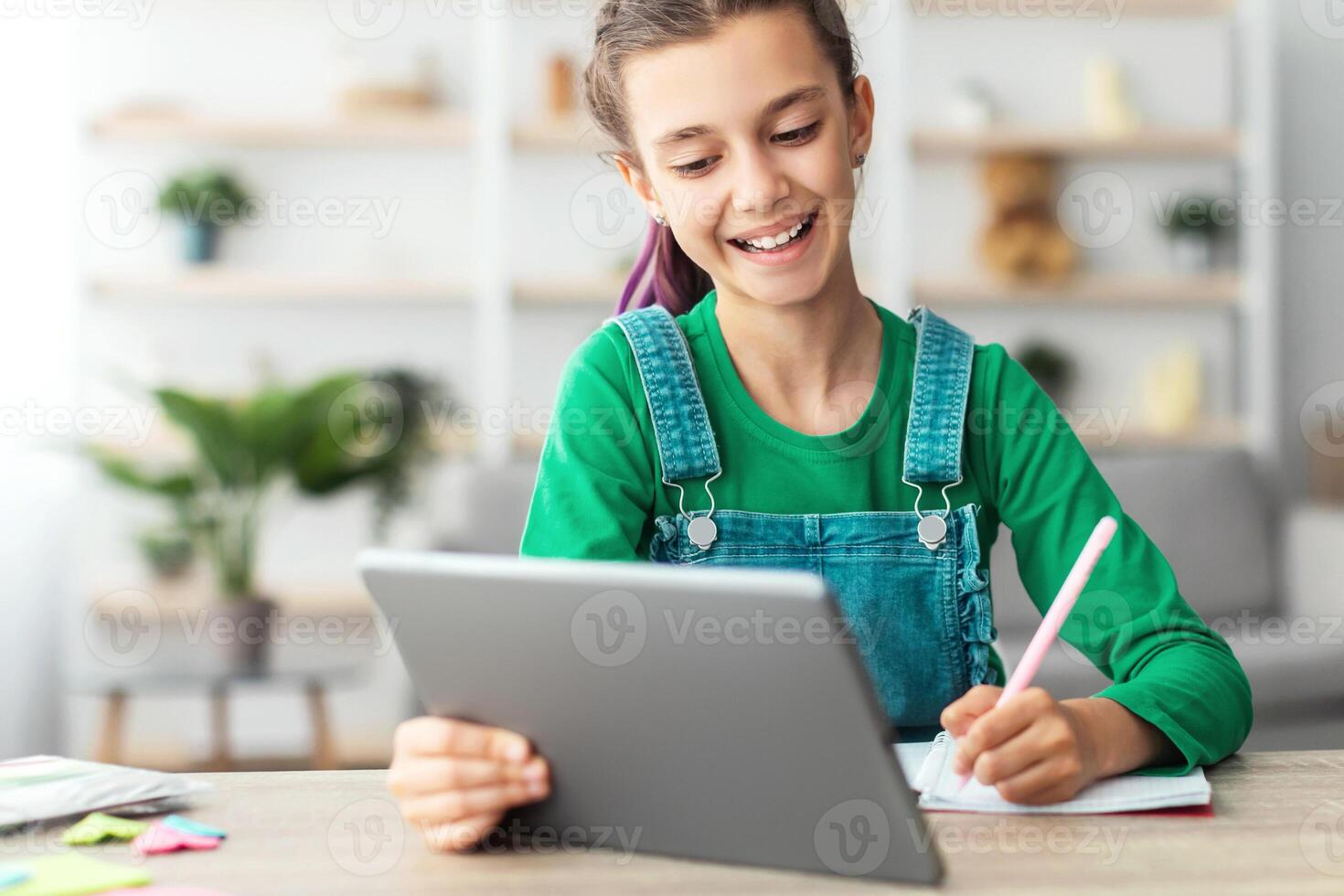 Remote Education Concept. Smiling young teenage girl sitting at desk, holding and using digital tablet writing in her notebook. Schoolgirl watching online course, taking notes, doing homework photo