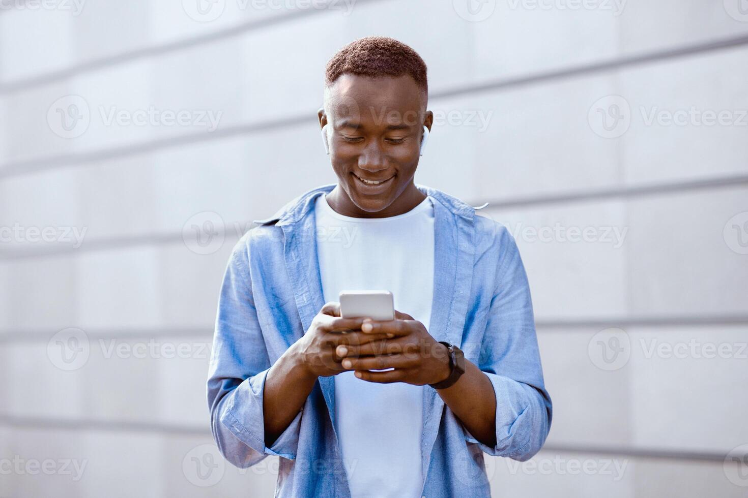 Cool African American man in earphones listening to music on cellphone near brick wall downtown. Funky black guy with mobile device choosing his favorite playlist or audio book outside photo