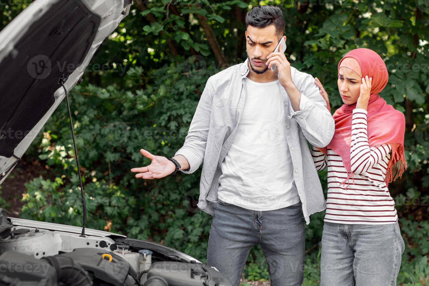 Sad despaired arab woman in hijab and man call to auto support, stand near broken car with open hood outdoor in summer, copy space. Problems on road, auto breakdown, family travel, vacation together photo