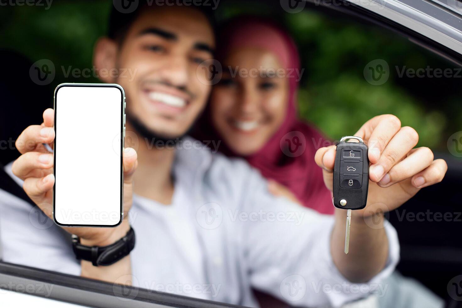 Car Rental App. Happy Muslim Couple Showing Key And Smartphone With Blank White Screen Through Window While Sitting In Automobile, Arab Spouses Recommending Travel Insurance, Closeup Shot, Mockup photo