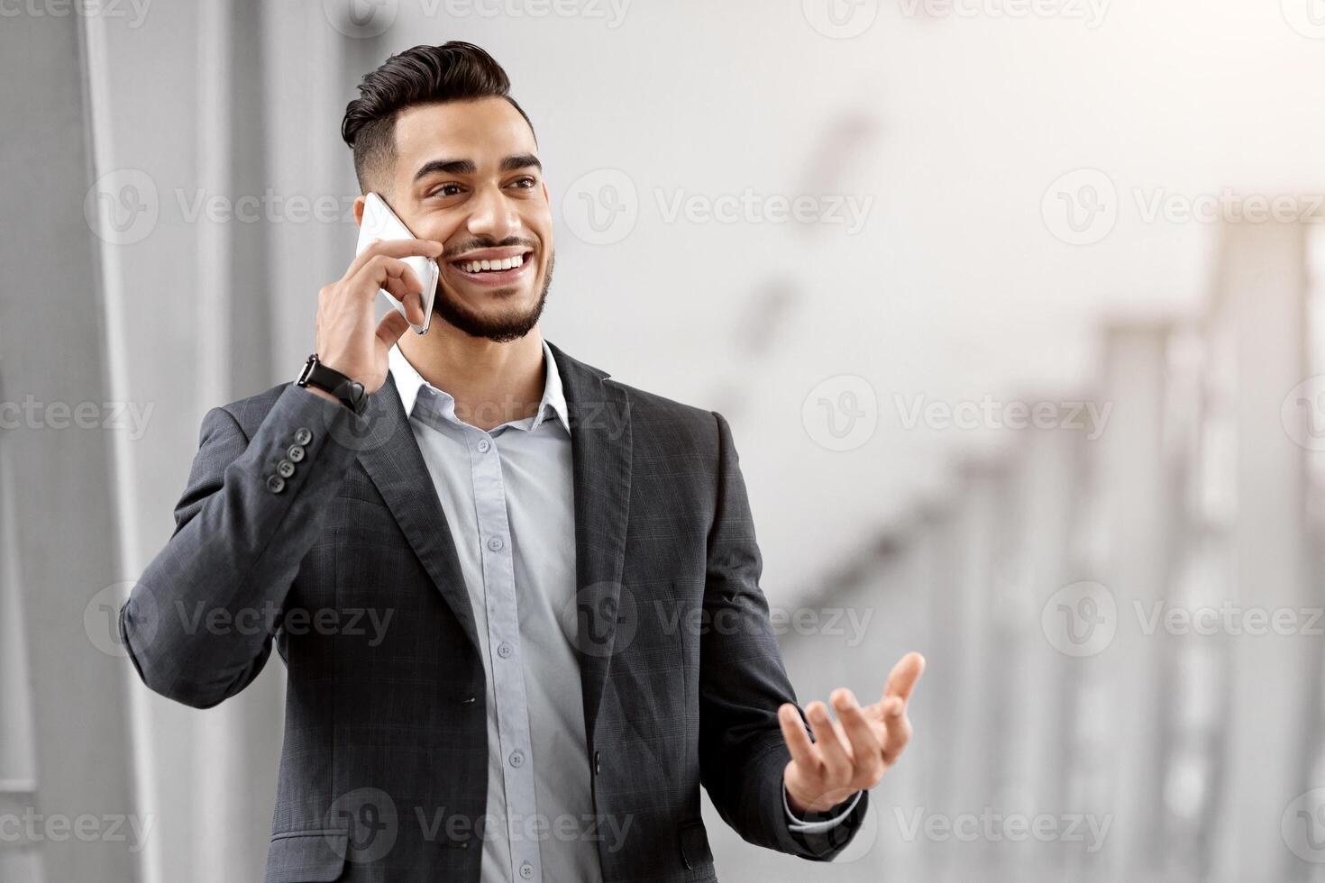 Handsome Arab Entrepreneur Talking On Mobile Phone While Waiting For Flight Departure At Airport, Smiling Middle Eastern Businessman In Suit Standing At Terminal And Enjoying Cellphone Conversation photo
