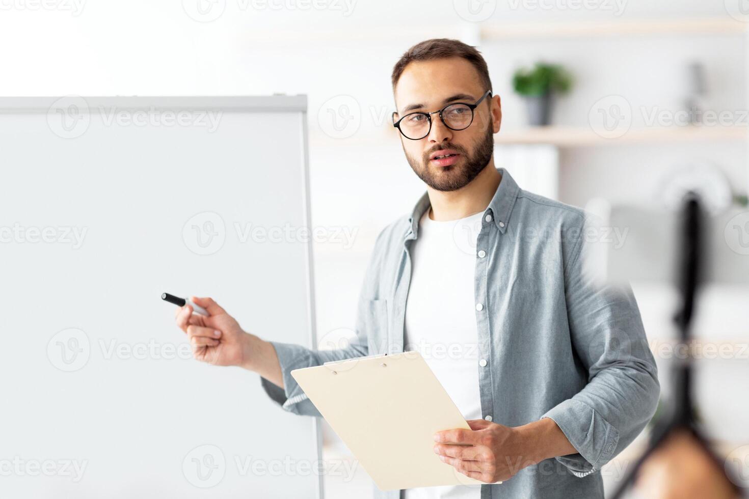 Young male teacher pointing at blackboard with English grammar rules, conducting online lesson from home. Caucasian tutor giving remote foreign language class, indoors photo
