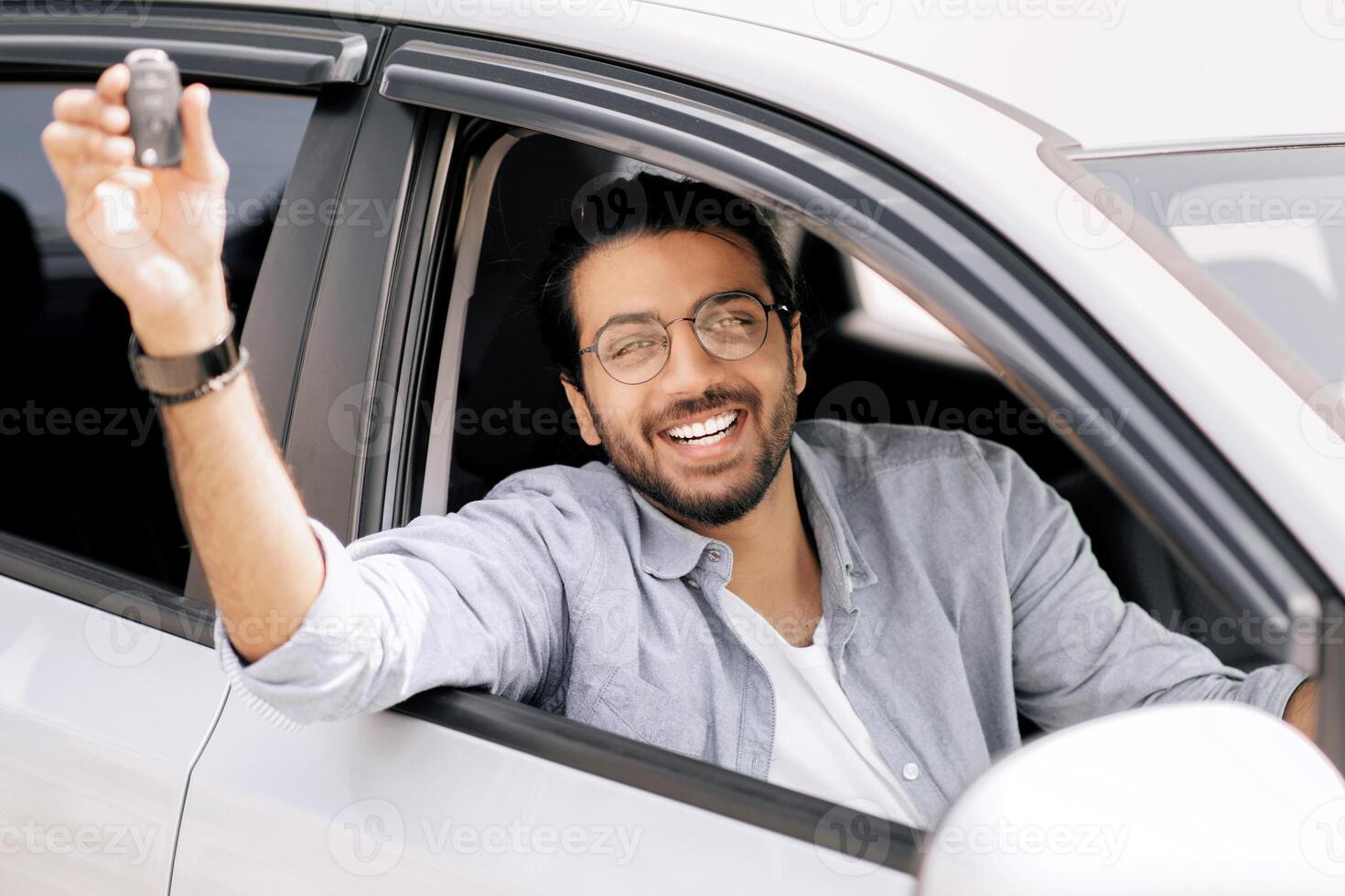 Cheerful indian long-haired guy with glasses sitting inside nice car and showing automatic key at camera, positive arab man renting automobile for travelling, having car ride or test-drive photo