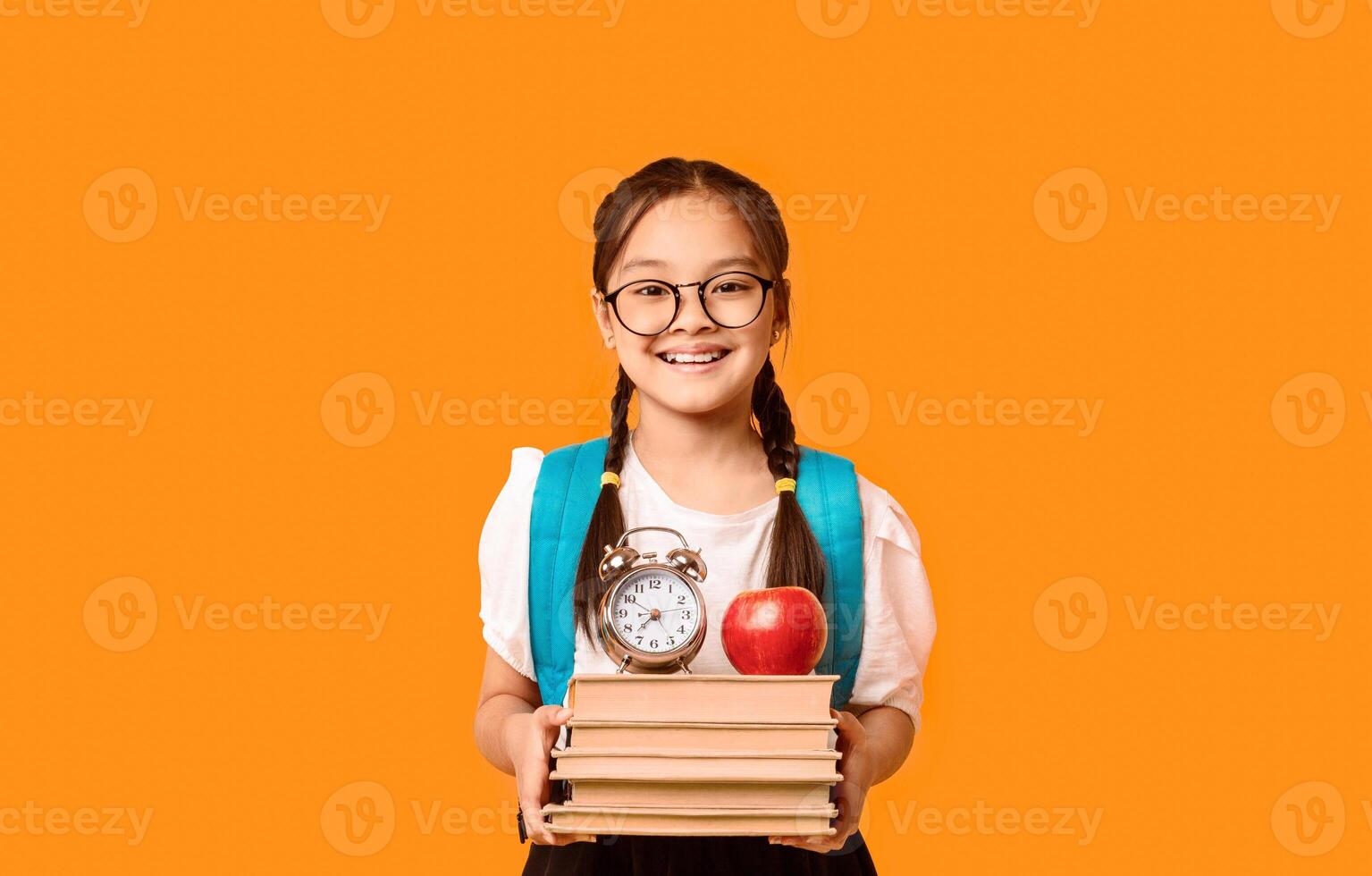 Back To School. Happy Chinese Schoolgirl With Backpack Holding Books Posing Standing In Studio On Yellow Background, Smiling To Camera photo