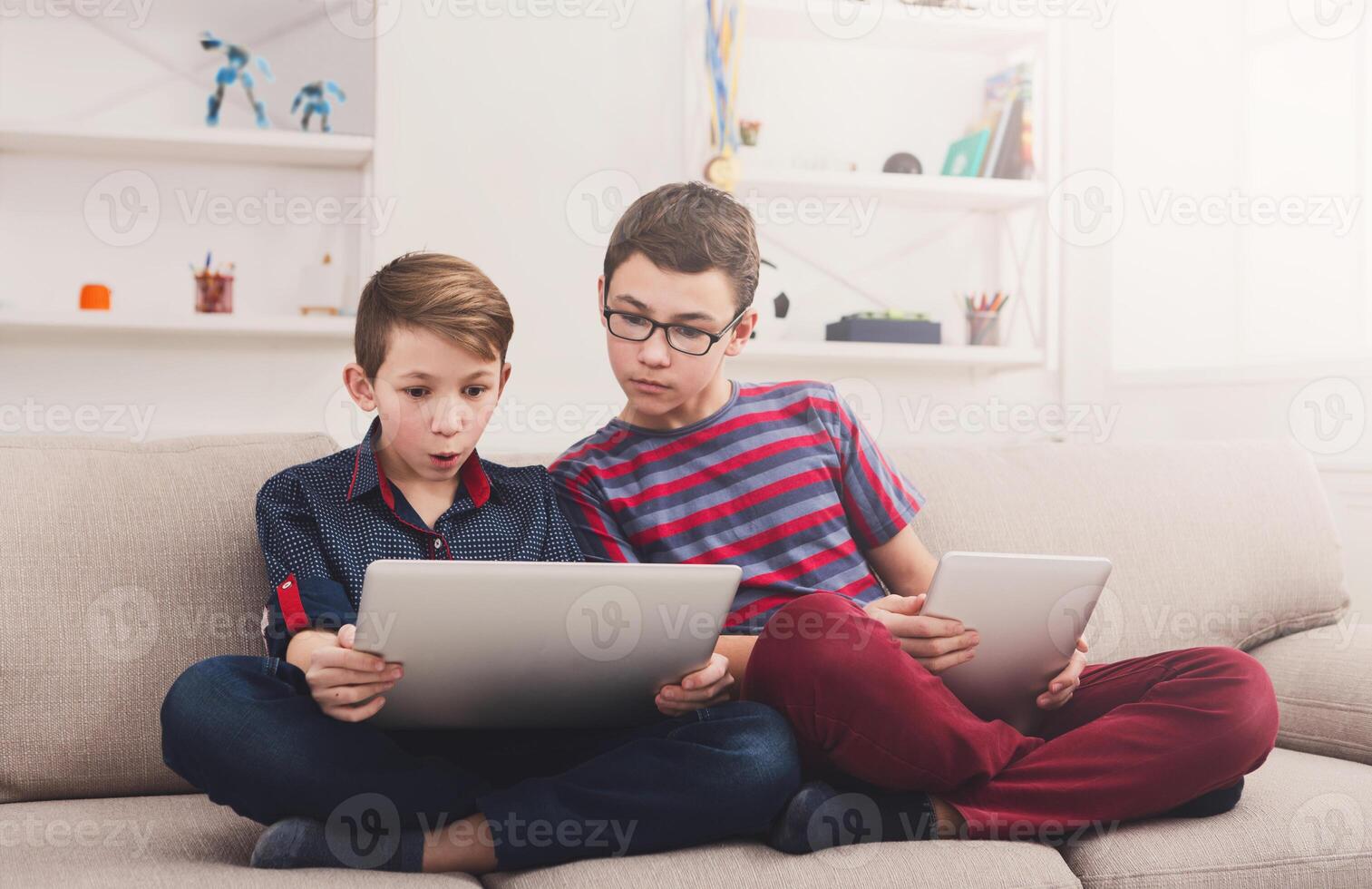 Two male kids with gadgets. Showing each other funny content on laptop and digital tablet on sofa at home. Boy friendship and communication concept photo