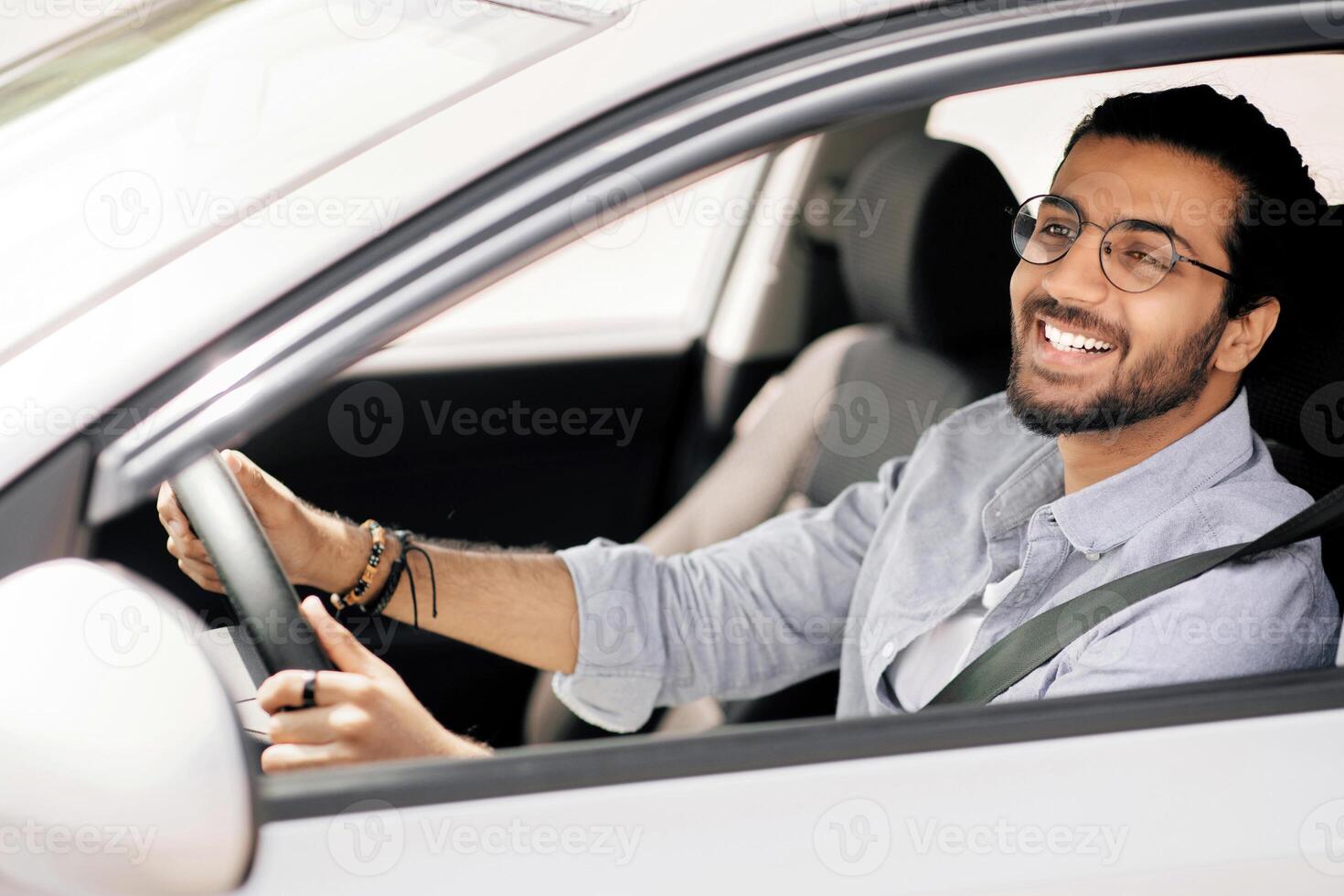 Happy millennial long-haired middle-eastern guy driving nice auto, wearing glasses, side view, shot from outside, copy space. Positive indian man holding hands on wheel, looking at road and smiling photo