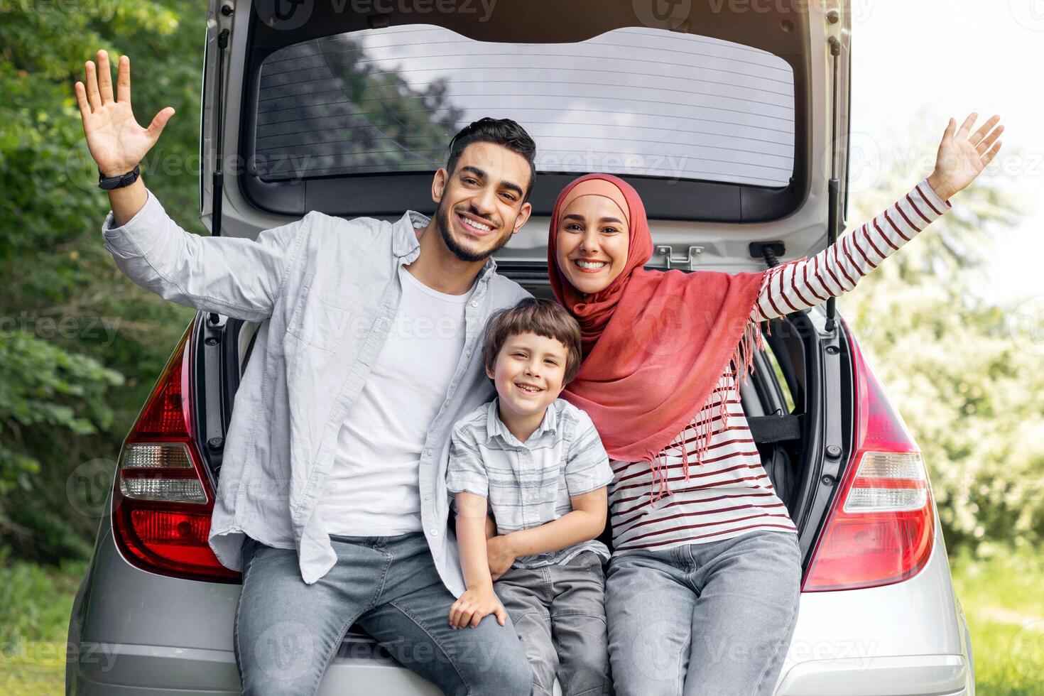 Satisfied millennial arab husband, wife in hijab and small boy waving hands sitting in car trunk, outdoors in summer, free space. Family weekend, vacation together and auto trip and life insurance photo