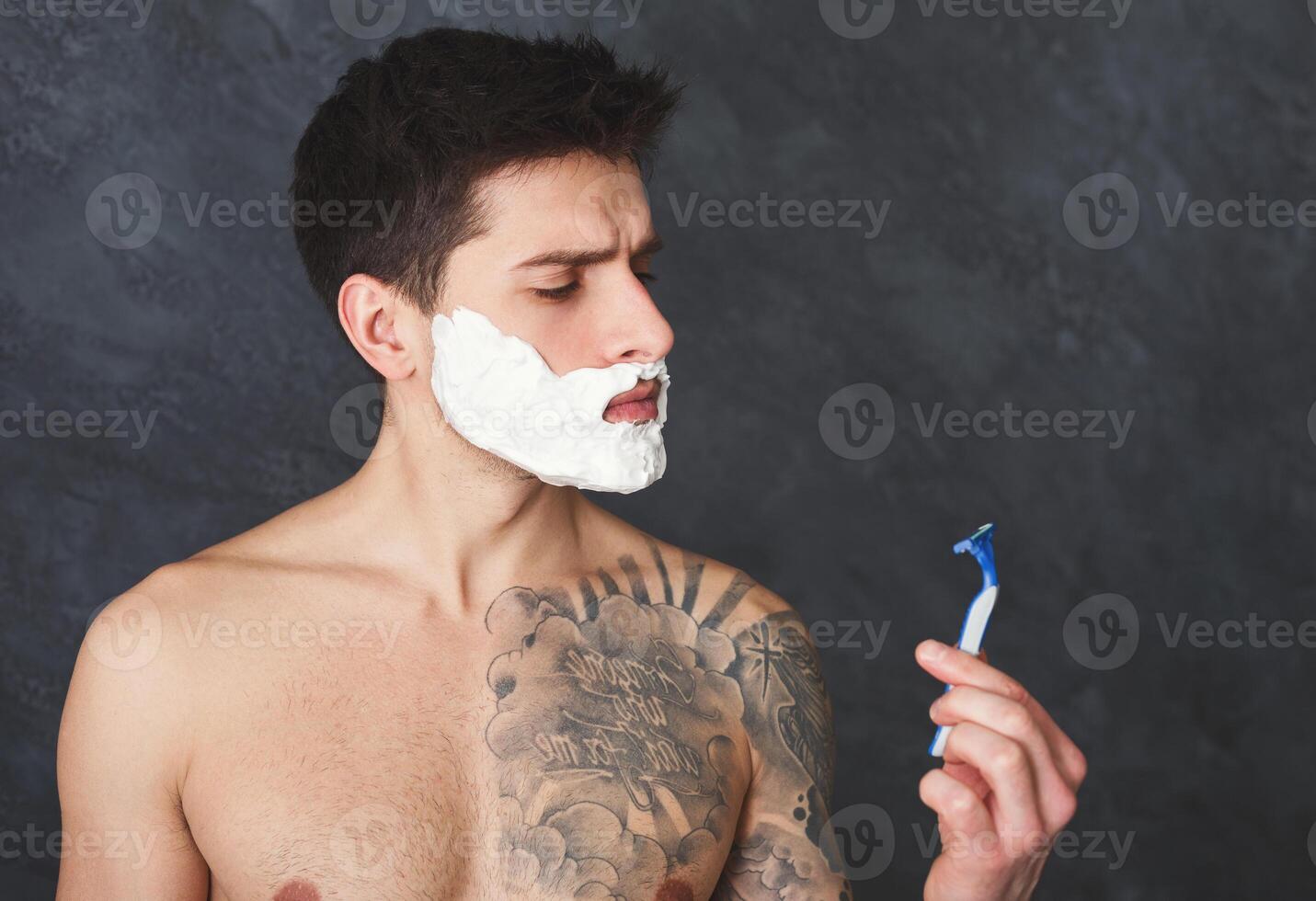 Young sexy tattooed man with foam on face looking at razor with blunt blade in hand. Topless guy with unpleased look shaving, gray studio background, copy space photo